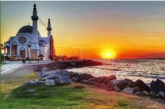 the sun is setting over an ocean with a church in the foreground and people walking on the shore