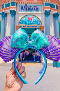 a hand holding up a purple and blue minnie mouse ears headband in front of a building