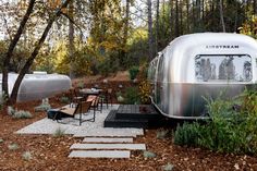 an airstream sits in the woods next to some chairs and tables with trees around it