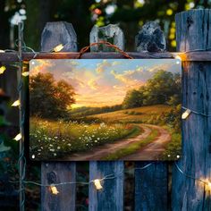 a painting hanging on a wooden fence with fairy lights strung around it and an image of a country road in the background