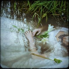 a woman laying down in the water with flowers on her arm and feet, holding onto a flower