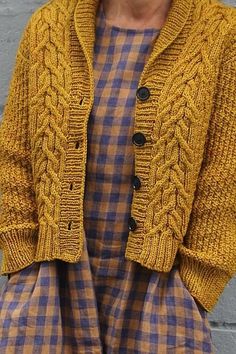 a woman standing in front of a gray wall wearing a yellow sweater and plaid pants