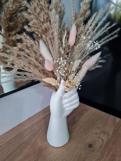 a white vase with dried flowers in it on a wooden table next to a mirror