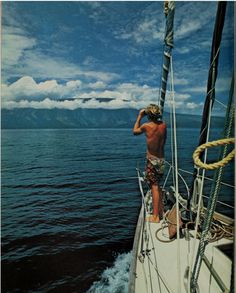 a man standing on the side of a boat looking out at the ocean with mountains in the background