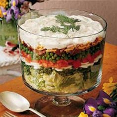 a layered salad in a glass dish on a wooden table with flowers and spoons