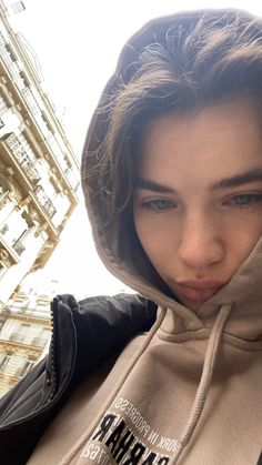 a young woman wearing a hoodie and looking at the camera while standing in front of a tall building