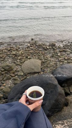 a person is holding a cup of tea on the rocks by the water's edge