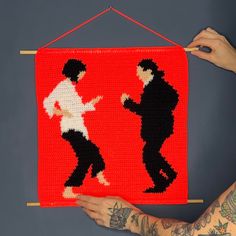 a person is holding up a crocheted wall hanging with two people on it