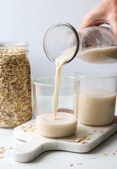 a person pouring milk into two glasses on top of a cutting board next to oatmeal