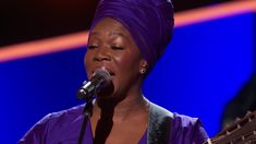 a woman with a purple turban on her head singing into a microphone and holding a guitar