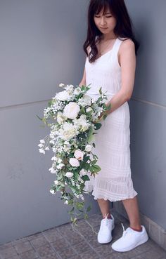 a woman holding a bouquet of flowers in front of a gray wall with white shoes