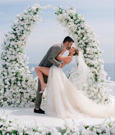 a bride and groom kissing in front of an arch of white flowers on the beach