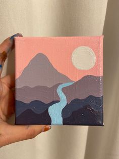 a person holding up a small painting with mountains and a river in the background,