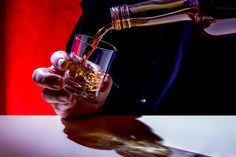 a person is pouring whiskey into a glass on a table with a red wall in the background