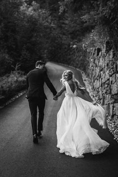 black and white photo of bride and groom holding hands walking down the road in front of a stone wall