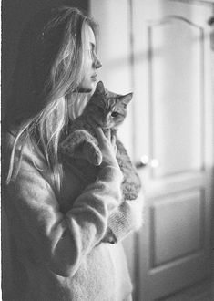 a woman holding a cat in her arms while standing next to a door and looking out the window