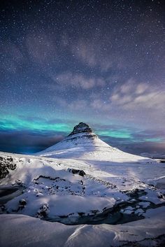 the sky is filled with green and blue lights as it stands on top of a snow covered mountain