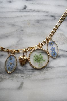 The Forget-Me-Not Meadow Charm Necklace is the ultimate summer accessory. It features real flowers, preserved in resin and pressed and encased in glass charms. Exact charms may vary slightly based on availability. The Forget-Me-Not Meadow Charm Necklace features: a single strand of 24K gold plated chain charm necklace with real preserved Queen Anne's lace, blue forget-me-not and rose bud flowers, and micro gold heart locket charms. Perfect for your long hot mornings on the beach with sandy toes Gold Flower Necklace With Vintage Charm, Gold Jewelry With Flower Charm Keepsake, Resin Round Pendant With Flower Charm, Gold Resin Flower Pendant Jewelry, Gold Flower-shaped Jewelry With Lobster Clasp, Vintage Gold Resin Jewelry, Gold Glass Flower Jewelry, Gold Flower-shaped Glass Jewelry, Gold Resin Necklace With Birth Flower Detail