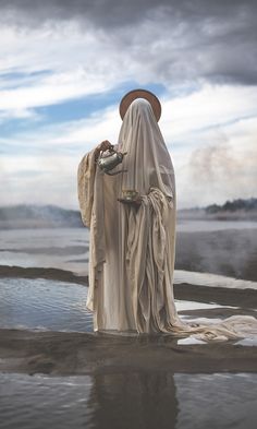 a woman dressed in white standing next to a body of water with a hat on her head