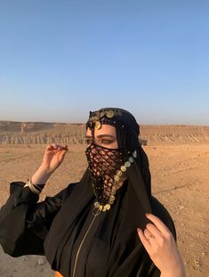 a woman wearing a black veil and headdress stands in the desert with her hands on her hips