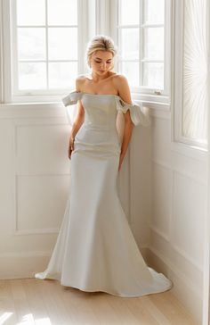 a woman standing in front of a window wearing a white wedding dress with off the shoulder sleeves