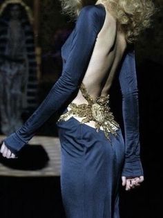 a woman with very long blonde hair walking down a runway wearing a blue dress and gold belt