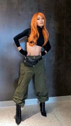 a woman with long red hair is standing in front of a wall wearing green cargo pants and black boots