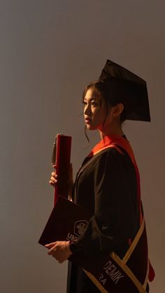 a woman wearing a graduation gown and holding a red book in one hand while standing against a white wall