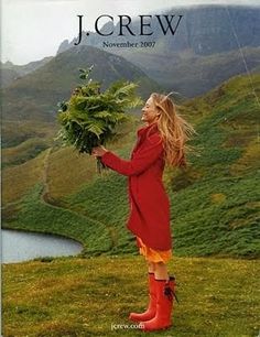 a woman in red coat and boots holding flowers on top of a green hill next to water