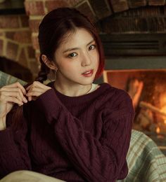 a young woman sitting in front of a fire place wearing a red sweater and earrings