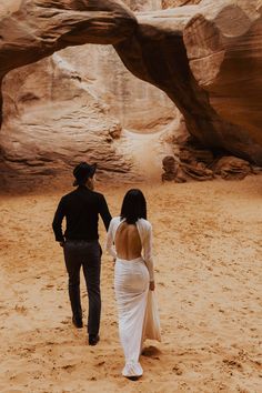 a man and woman are walking through the desert with an arch in the back ground