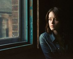 a woman sitting in front of a window next to a brick wall and looking into the distance