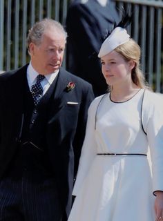 an older man and young woman dressed in formal attire walking together outside on a sunny day