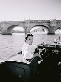 black and white photograph of man driving boat on river with bridge in the back ground