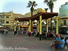 people are sitting on benches and tables in the middle of a plaza with palm trees