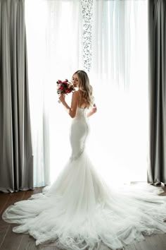 a woman standing in front of a window wearing a wedding dress and holding a bouquet