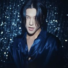 a man with black hair wearing a blue shirt in front of a shiny wall and looking at the camera