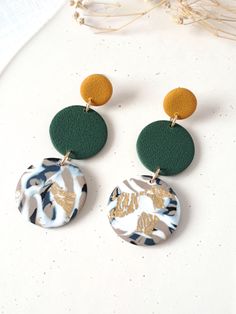 two circular earrings with green and gold accents on top of a white table next to dried flowers