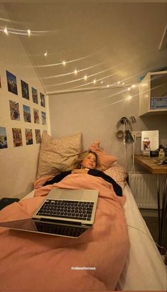 a woman laying in bed with a laptop on her lap and lights strung across the room