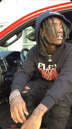 a man with dreadlocks sitting in the drivers seat of a car wearing a black hoodie