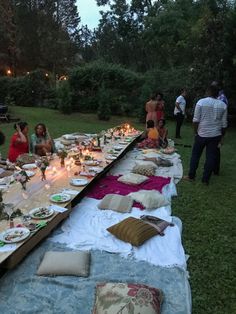 a group of people sitting at a long table with plates and candles in the grass