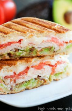 a grilled sandwich on a white plate with an avocado and tomato in the background