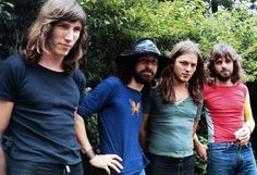 four young men standing next to each other in front of trees and bushes, one with long hair