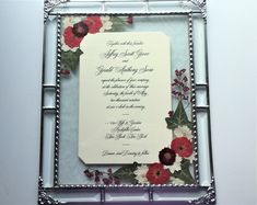 a wedding card with red and white flowers on it in a silver frame hanging from the wall