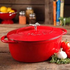 a red casserole sitting on top of a wooden table next to some vegetables