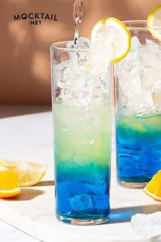 two glasses filled with blue and green liquid next to lemon wedges on a table