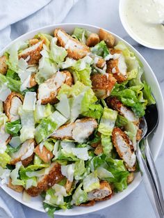 a white bowl filled with lettuce and chicken next to a fork on top of a table