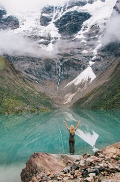 a woman standing on top of a rocky cliff next to a lake with snow covered mountains in the background