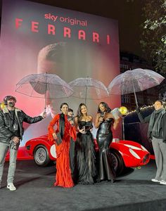 four people standing in front of a red car with umbrellas over their heads and one person holding an umbrella