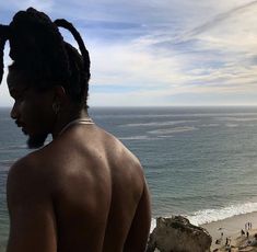 a man with dreadlocks looking out over the ocean from a cliff overlooking a beach